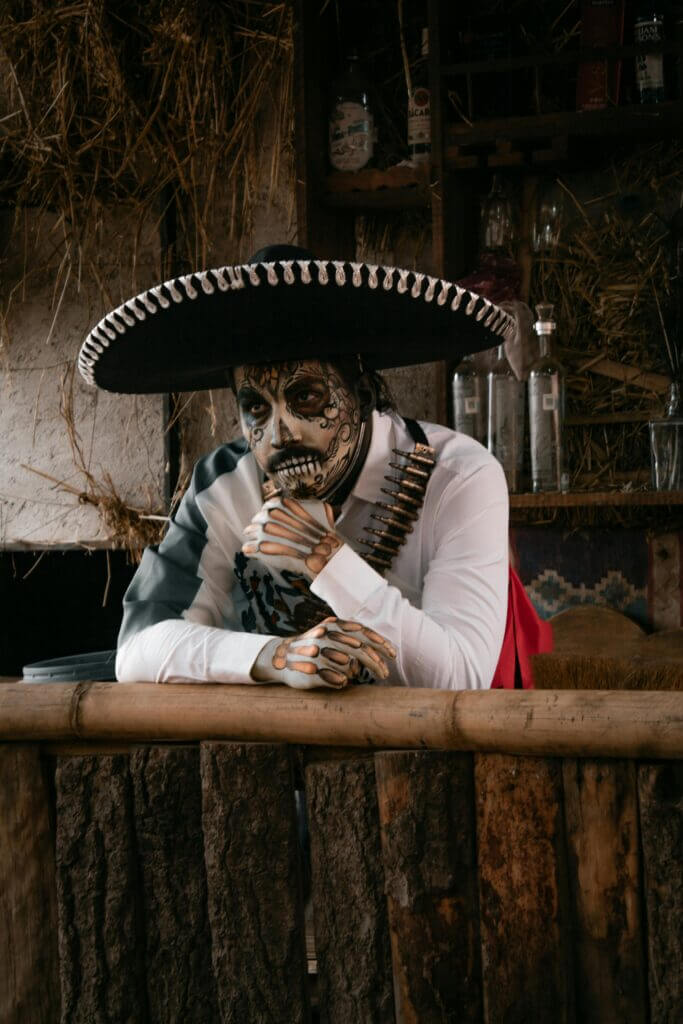 Portrait of a man in a Mexican costume with face paint, leaning on a wooden bar.